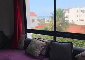 Una habitación con un ventanal grande, un sofá morado con cojines florales y vistas a un edificio de techo rojo y palmeras.
