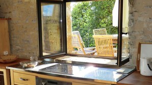 Une cuisine avec une grande fenêtre, des chaises en bois à l’extérieur et une machine à café posée sur le plan de travail.