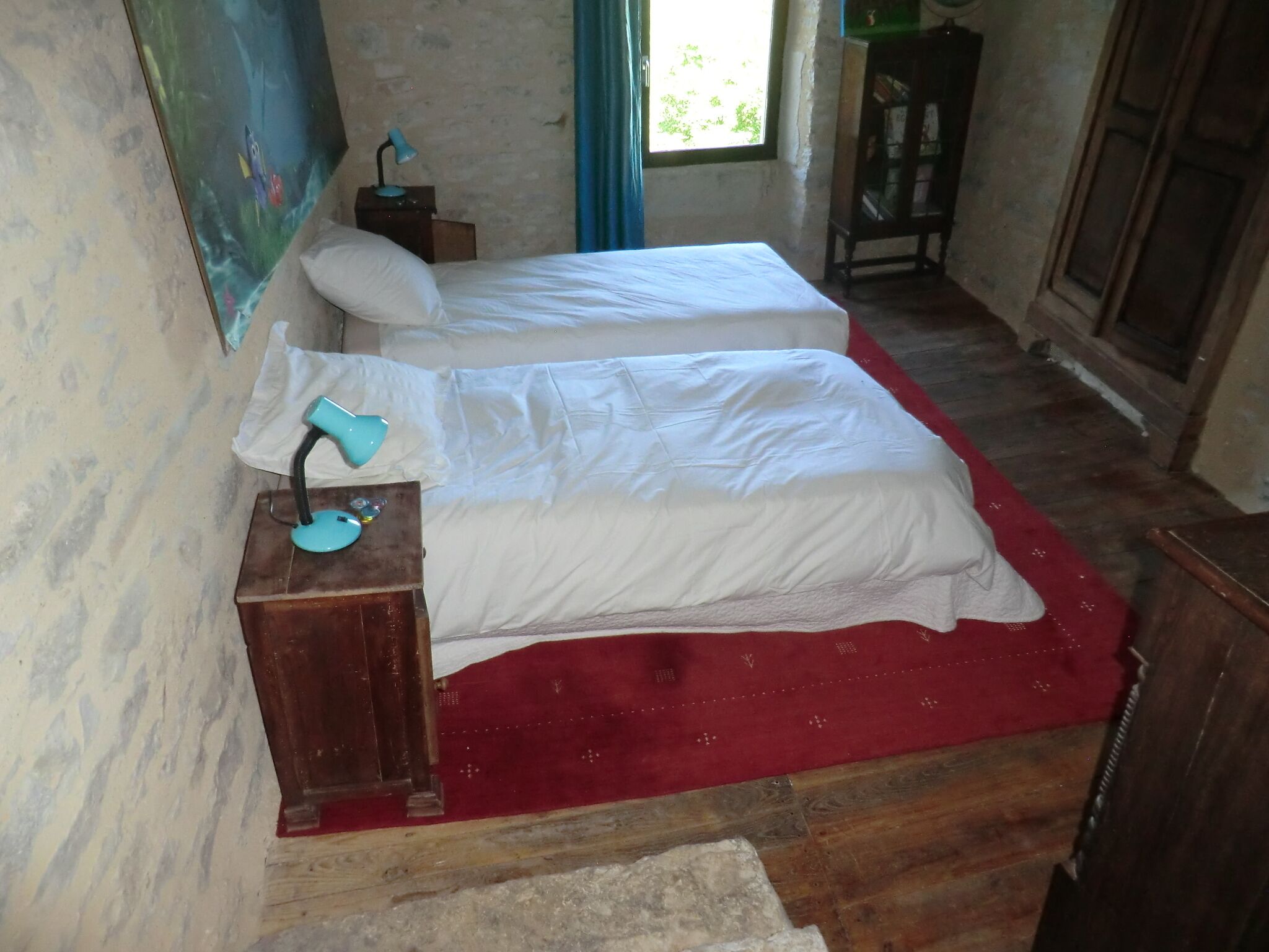 A room with two beds, a red rug, a small table with a lamp, and a painting on the wall.