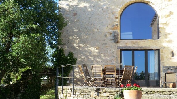 Un bâtiment en pierre doté d’une grande fenêtre en arc de cercle, d’un balcon avec des chaises en bois et d’une plante en pot.