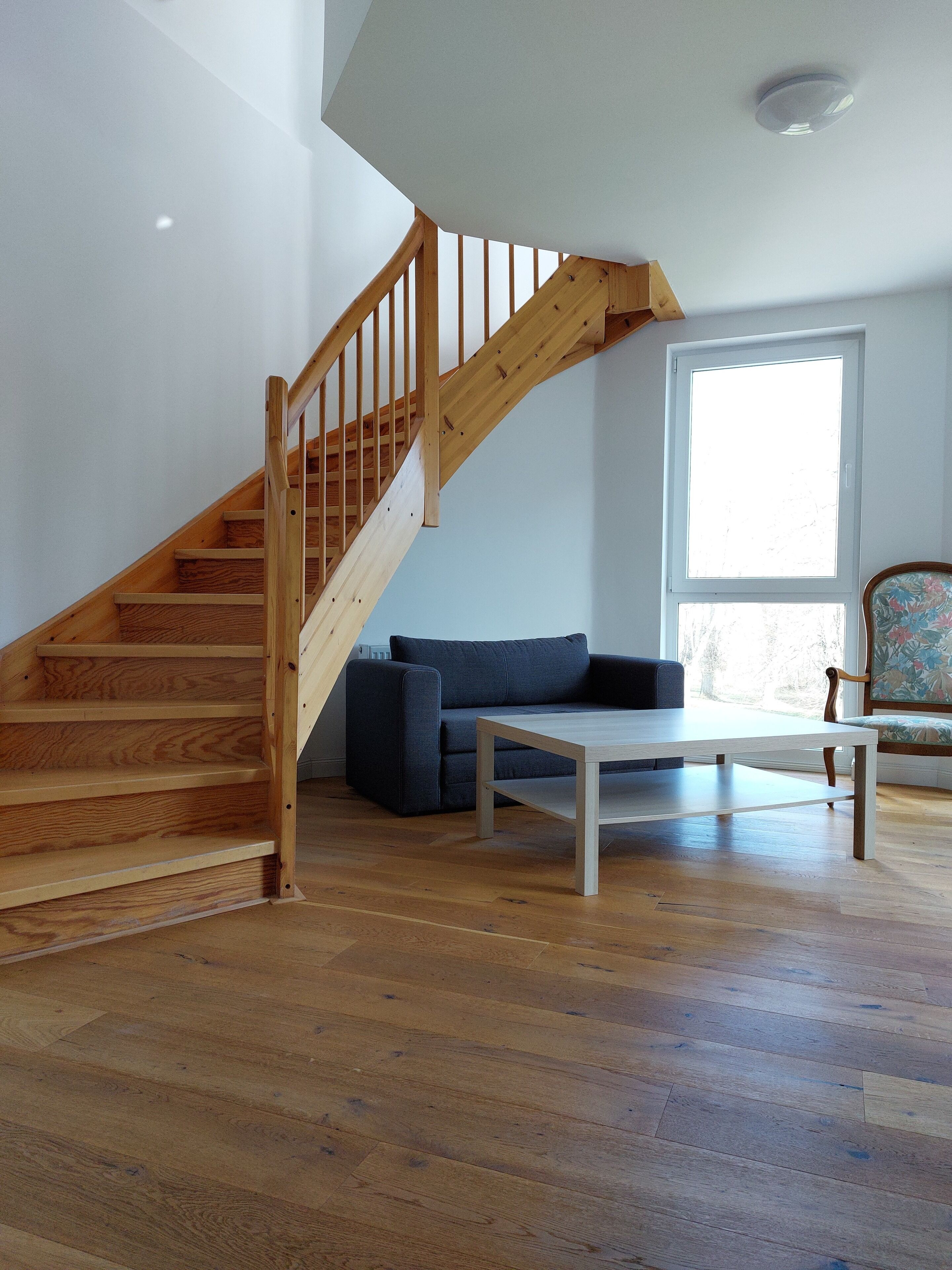 A wooden staircase leading to a second floor, a dark blue sofa, a white coffee table, and a window with a view of trees.