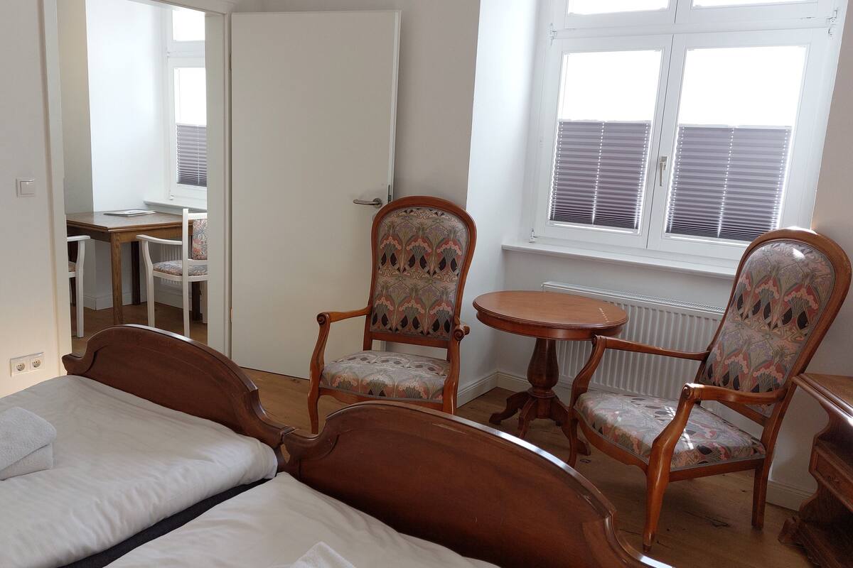 A bedroom with a bed, two wooden chairs, a small table, and a window with blinds.