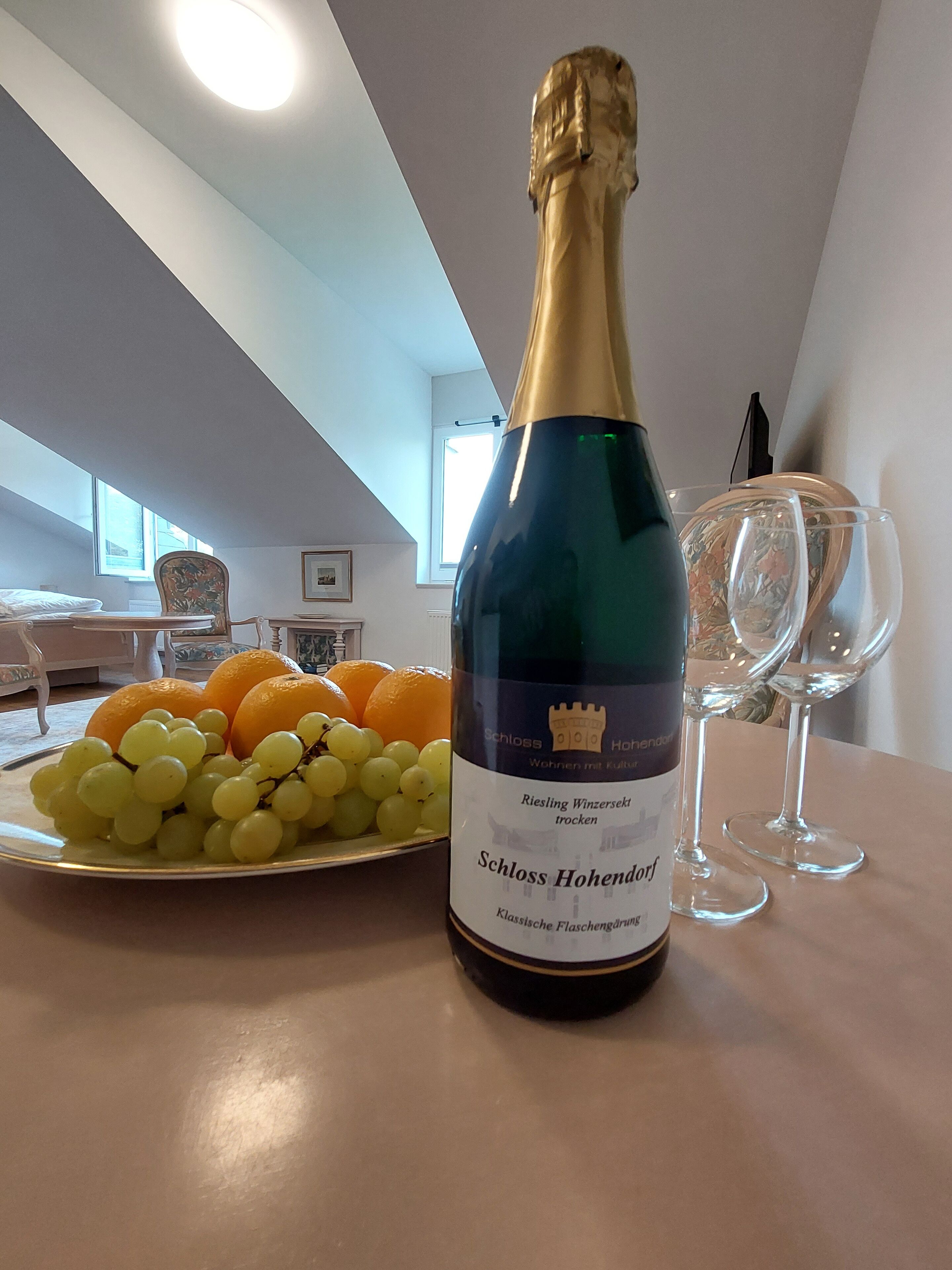Une bouteille de vin Riesling de la marque Schloss Hohendorf, une assiette de raisins et d’oranges, et deux verres à vin vides sur une table dans une pièce dont le plafond est en pente et qui possède une fenêtre.