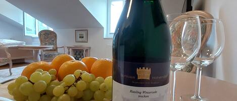 Pullo Schloss Hohendorf -rieslingviiniä, lautanen täynnä rypäleitä ja appelsiineja sekä kaksi tyhjää viinilasia pöydällä huoneessa, jossa on kalteva katto ja ikkuna.