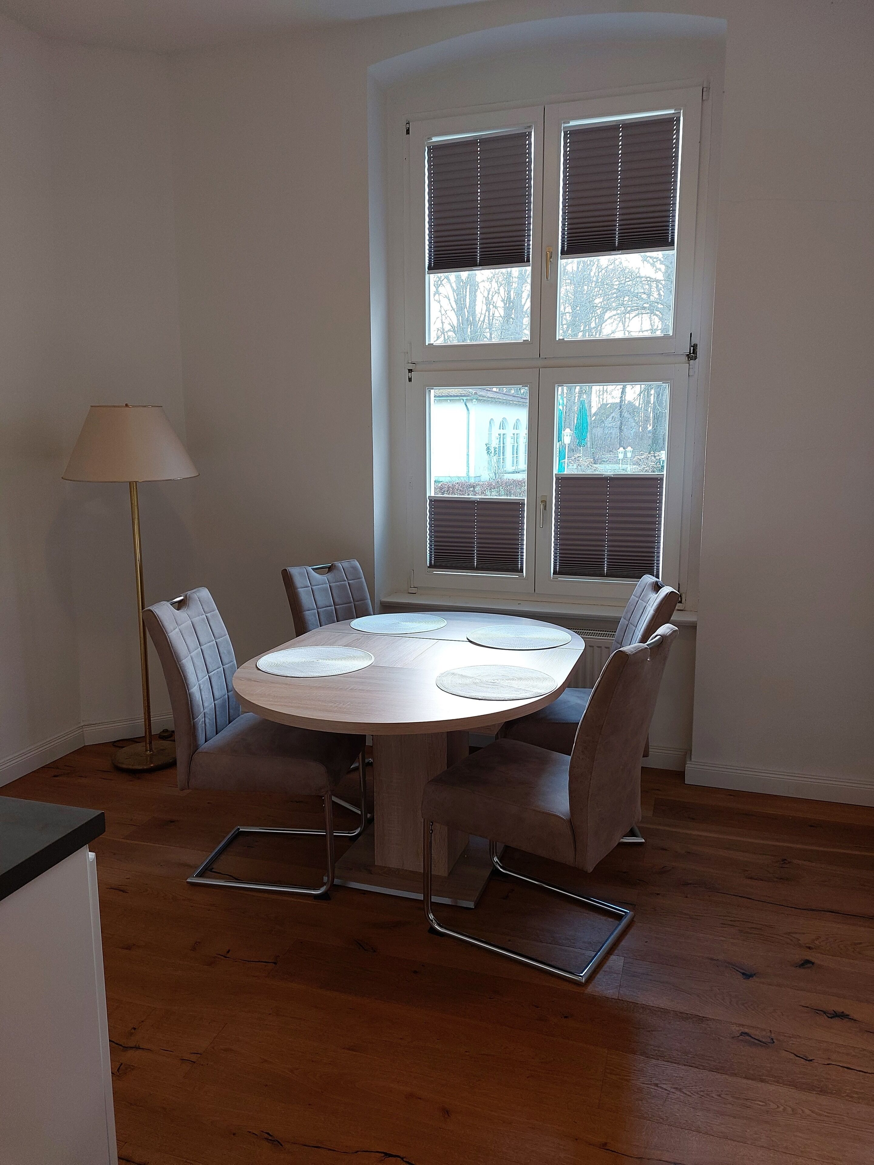 Un área de comedor con una mesa redonda, seis sillas, una lámpara de pie y un amplio ventanal con persianas.