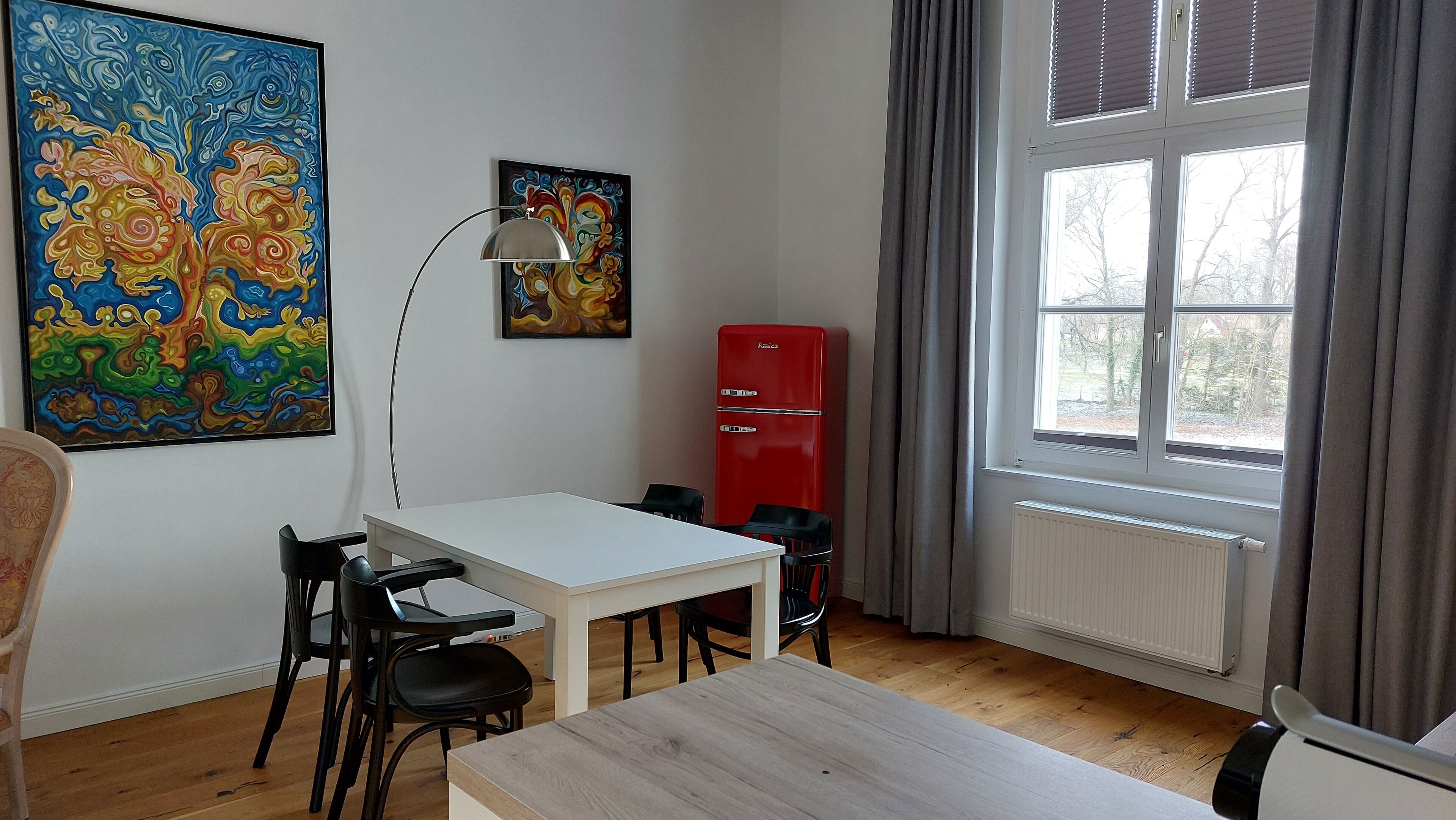 Una habitació amb una taula i cadires blanques, un refrigerador vermell, un quadre a la paret i una finestra amb cortines.