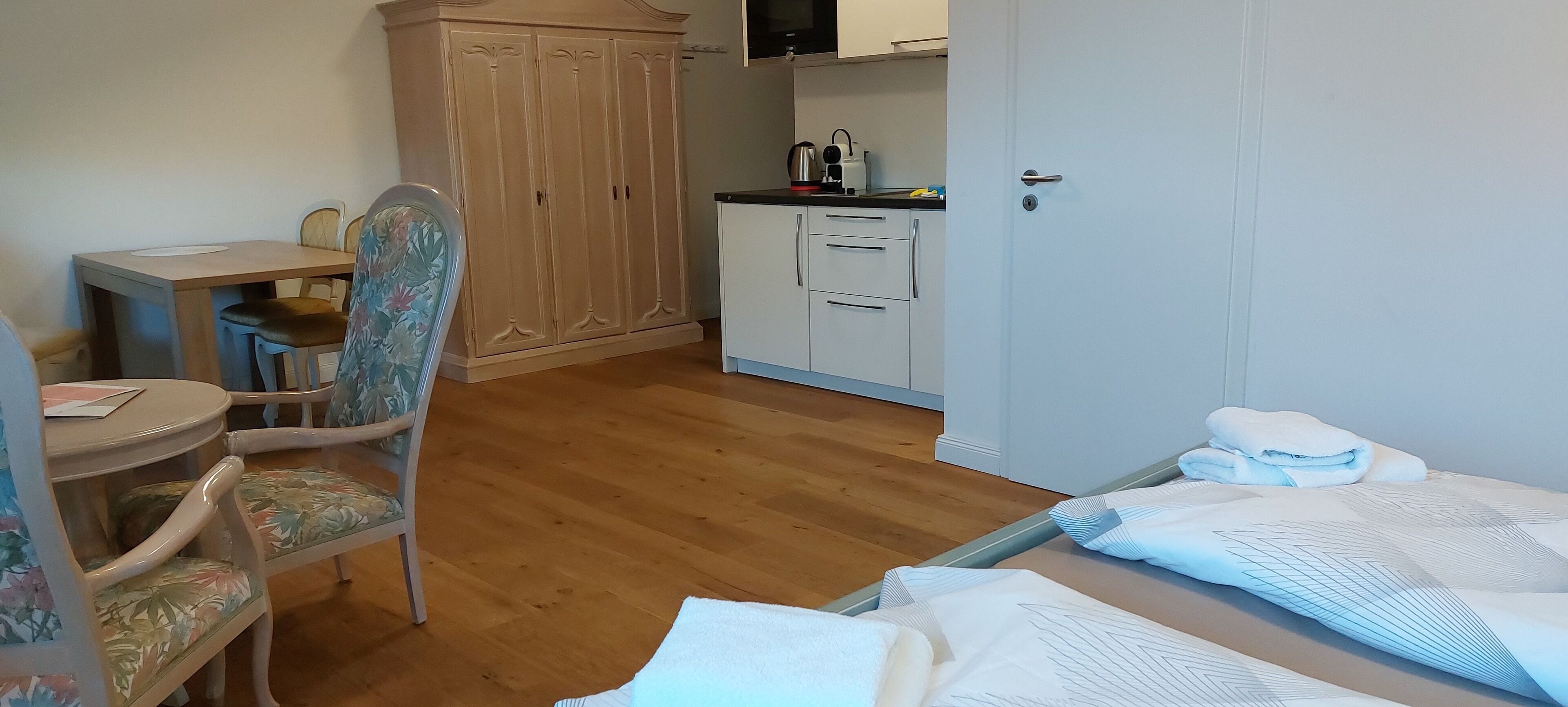 A room with a wooden floor, a dining table with chairs, a wardrobe, a kitchen area with cabinets and a microwave, and a bed with a white bedspread.