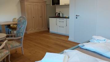 A room with a wooden floor, a dining table with chairs, a wardrobe, a kitchen area with cabinets and a microwave, and a bed with a white bedspread.