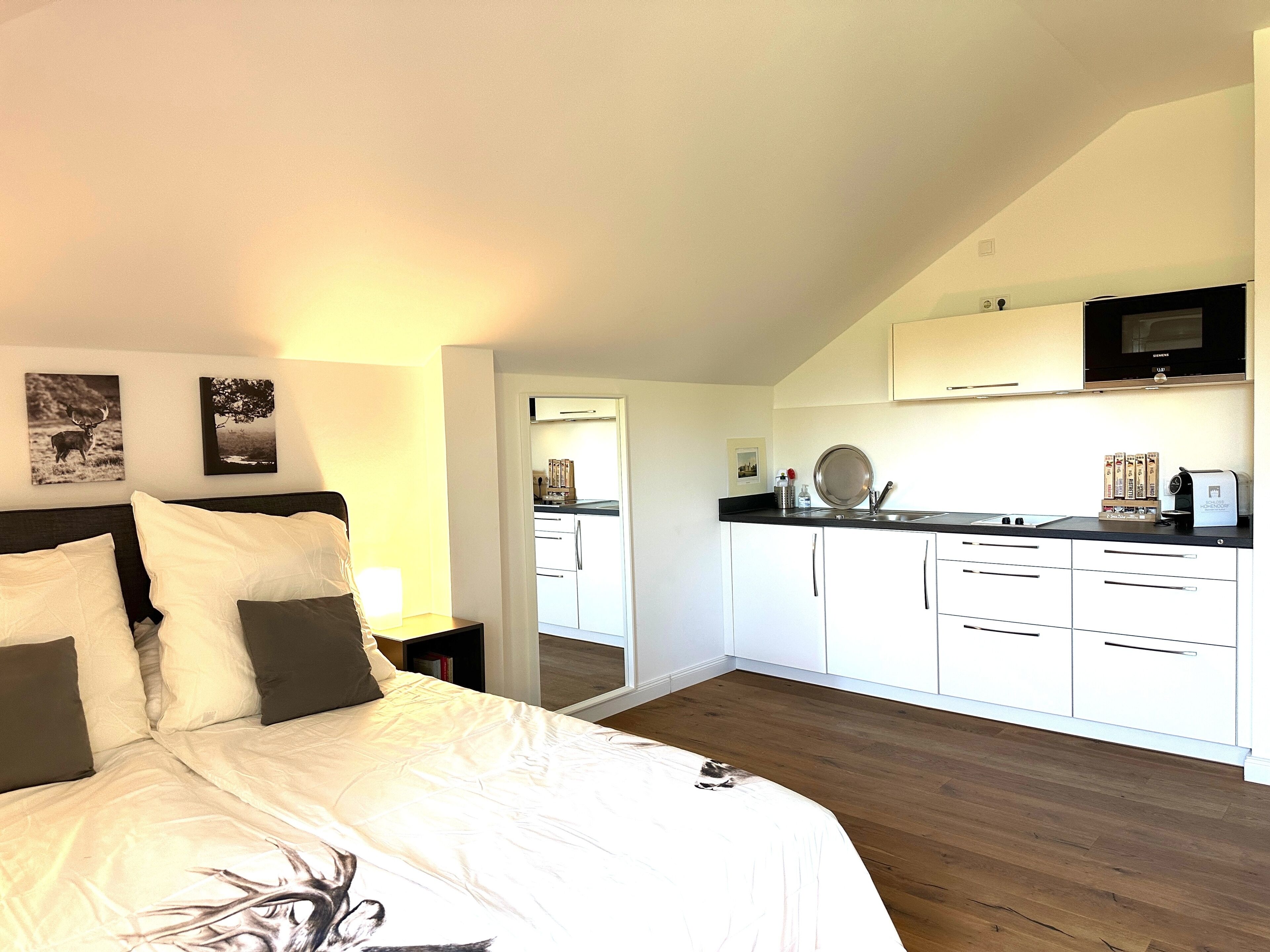 Een slaapkamer met een bed, een nachtkastje en een aan de muur bevestigde televisie. Grenzend aan de slaapkamer bevindt zich een keuken met witte kasten, een zwart aanrecht en een magnetron.