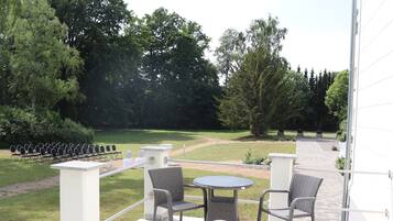 Un patio exterior con sillas de mimbre y una pequeña mesa redonda, con vistas a un área verde con filas de sillas y árboles.
