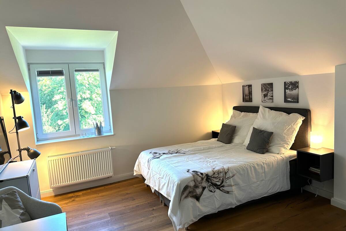 A bedroom with a large bed, a window with blinds, a radiator, and a desk with a lamp.