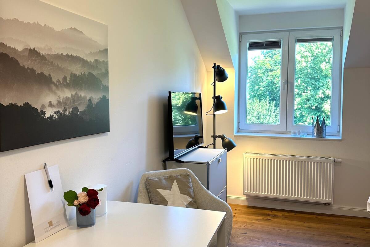 A modern room with a white table, a chair, a desk with a computer and a TV, a window with a view of trees, and a painting on the wall.