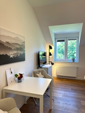 A modern room with a white table, a chair, a desk with a computer and a TV, a window with a view of trees, and a painting on the wall. - Schloss Hohendorf (Gross Mohrdorf)