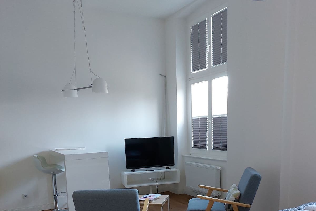 A modern hotel room with a bed, two chairs, a desk, a TV, and a large window.
