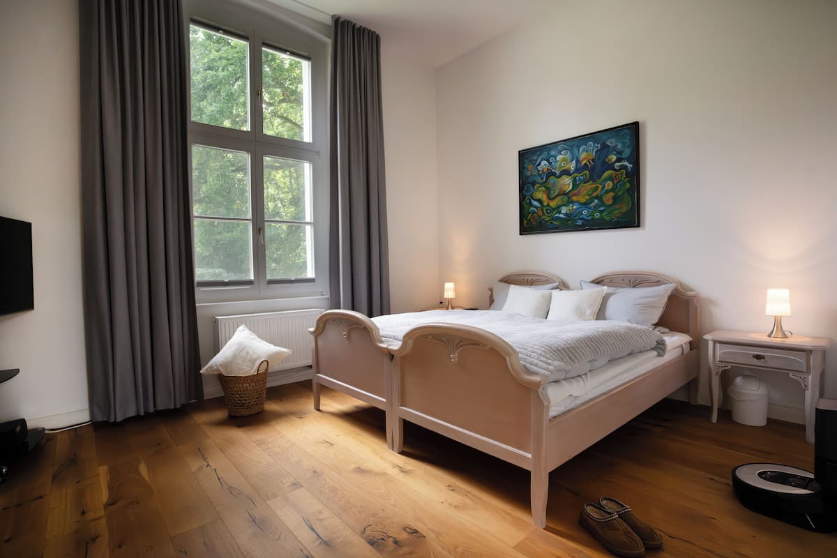 A bedroom with a double bed, a bedside table with a lamp, a radiator, a window with curtains, and a painting on the wall.