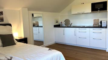 Un dormitorio con cama, mesita de noche y vistas a una cocina con armarios y microondas.