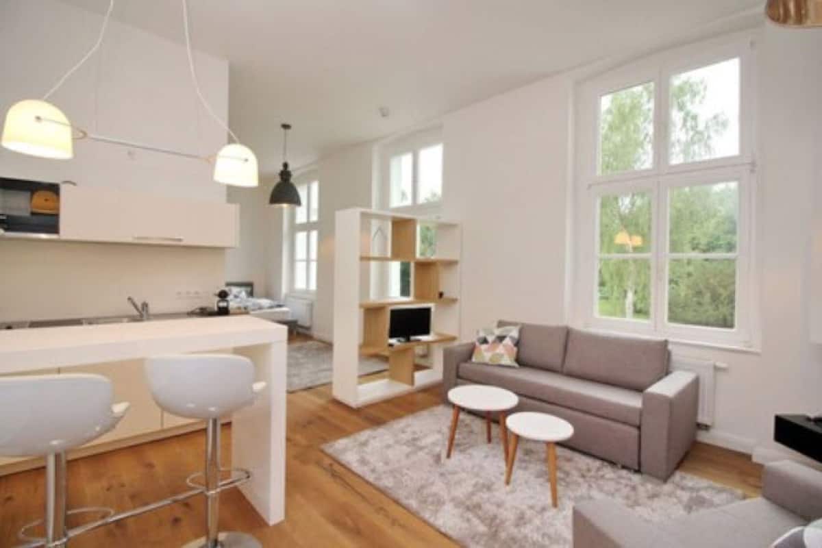 A modern kitchen with white cabinets, a bar area with stools, a grey sofa, and a TV on a wooden stand.