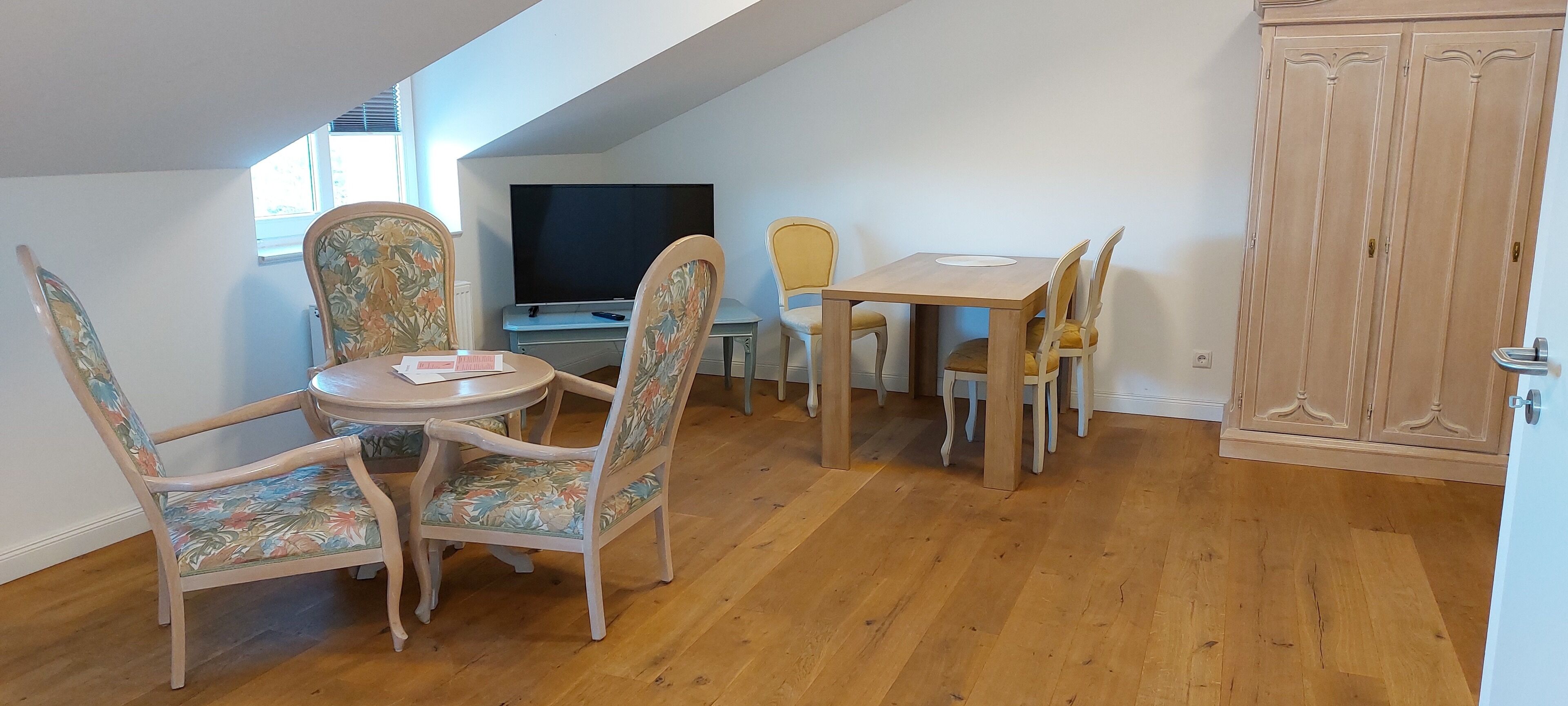 A dining area with a round table and floral chairs, a television on a stand, and wooden flooring.