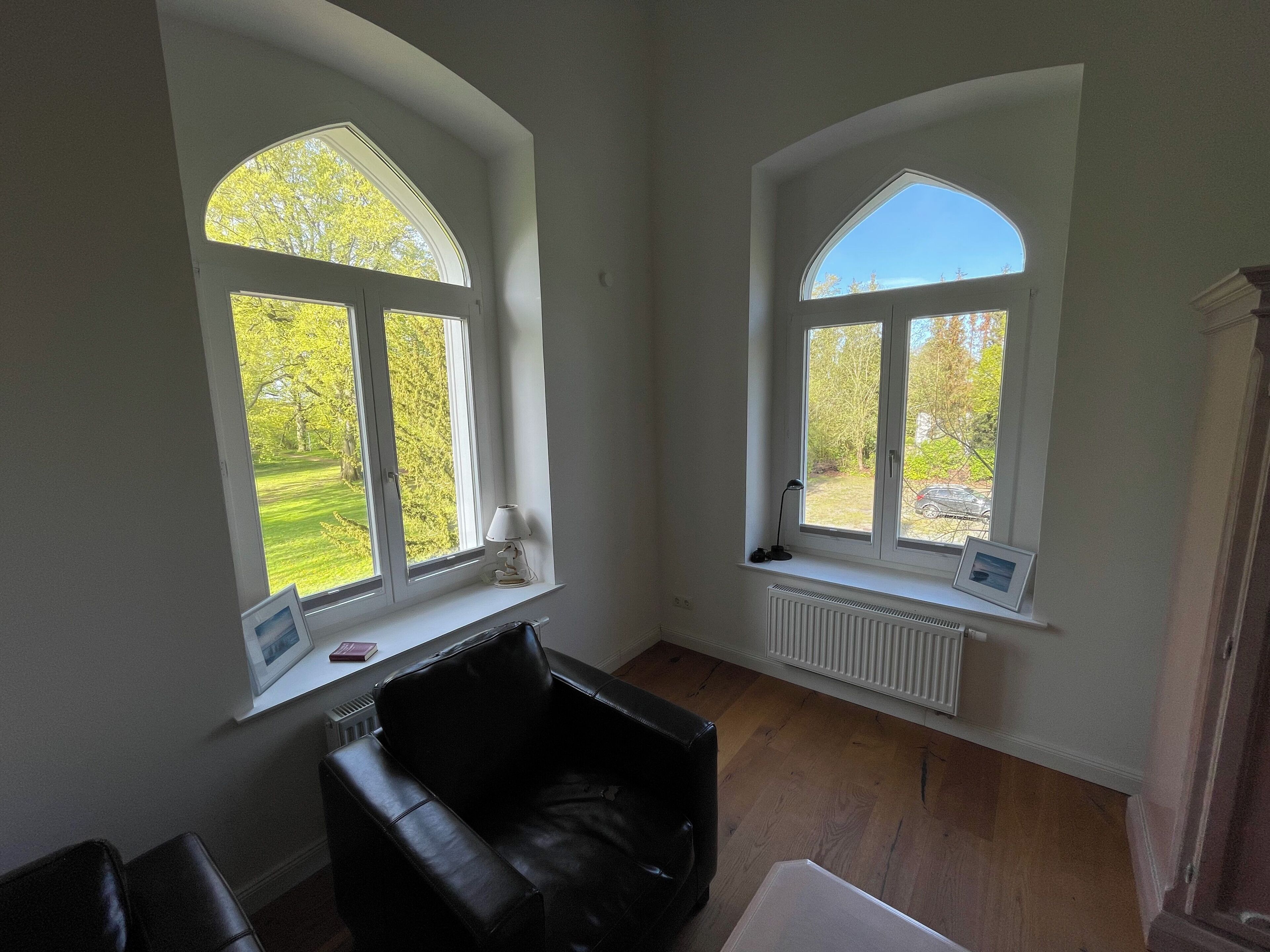 Ein Zimmer mit zwei Bogenfenstern, einem Heizkörper, einem schwarzen Ledersessel und einem weißen Tisch.
