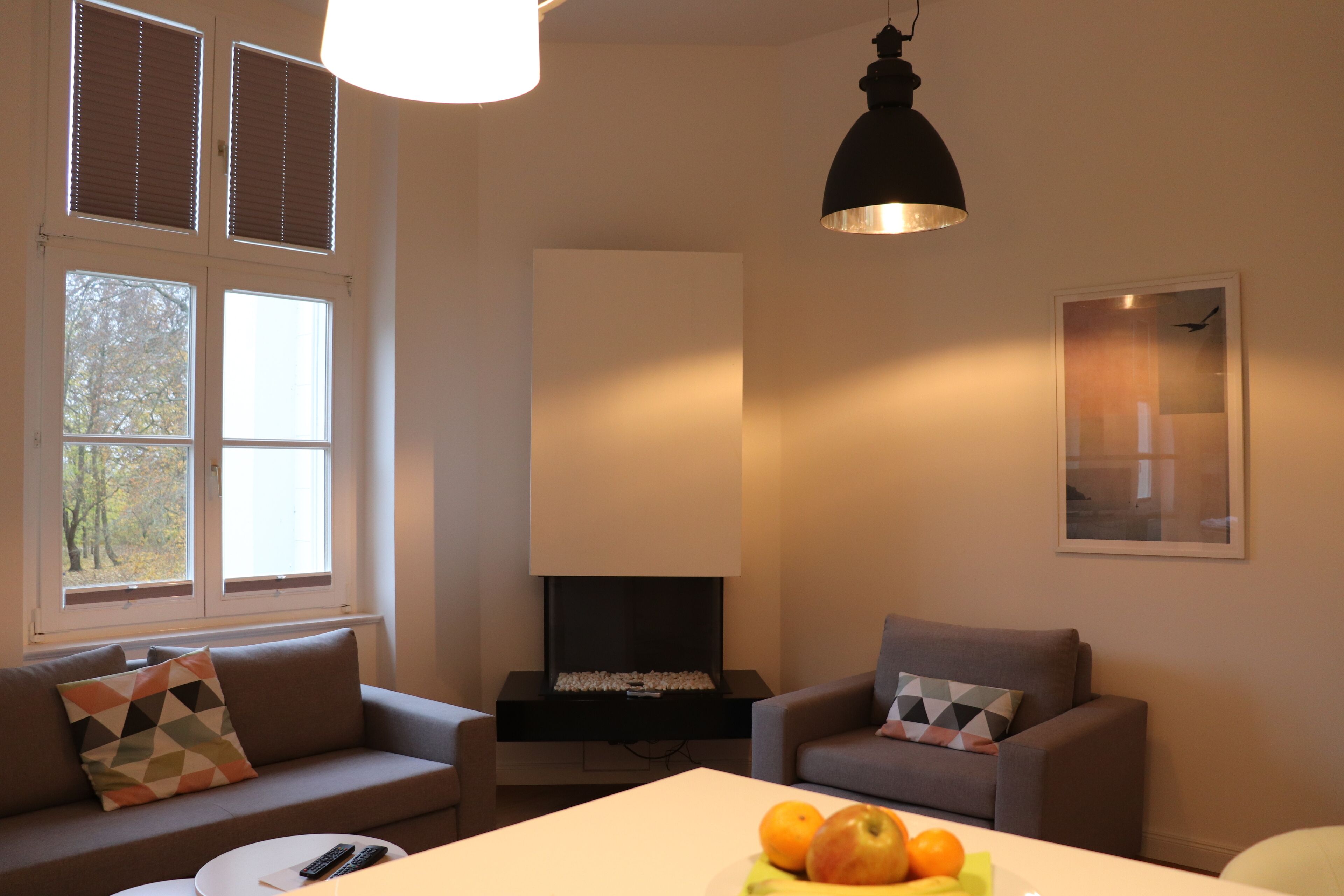 Una sala de estar con sofá, sillón y una mesa con fruta. Hay ventanas con persianas, una chimenea y una lámpara colgante.