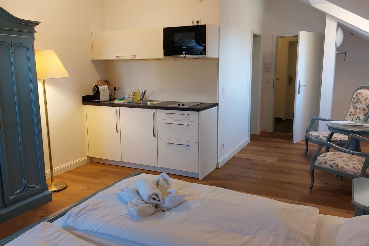 A compact hotel room with a kitchenette, a bed with towels, a desk with a chair, and a wardrobe.