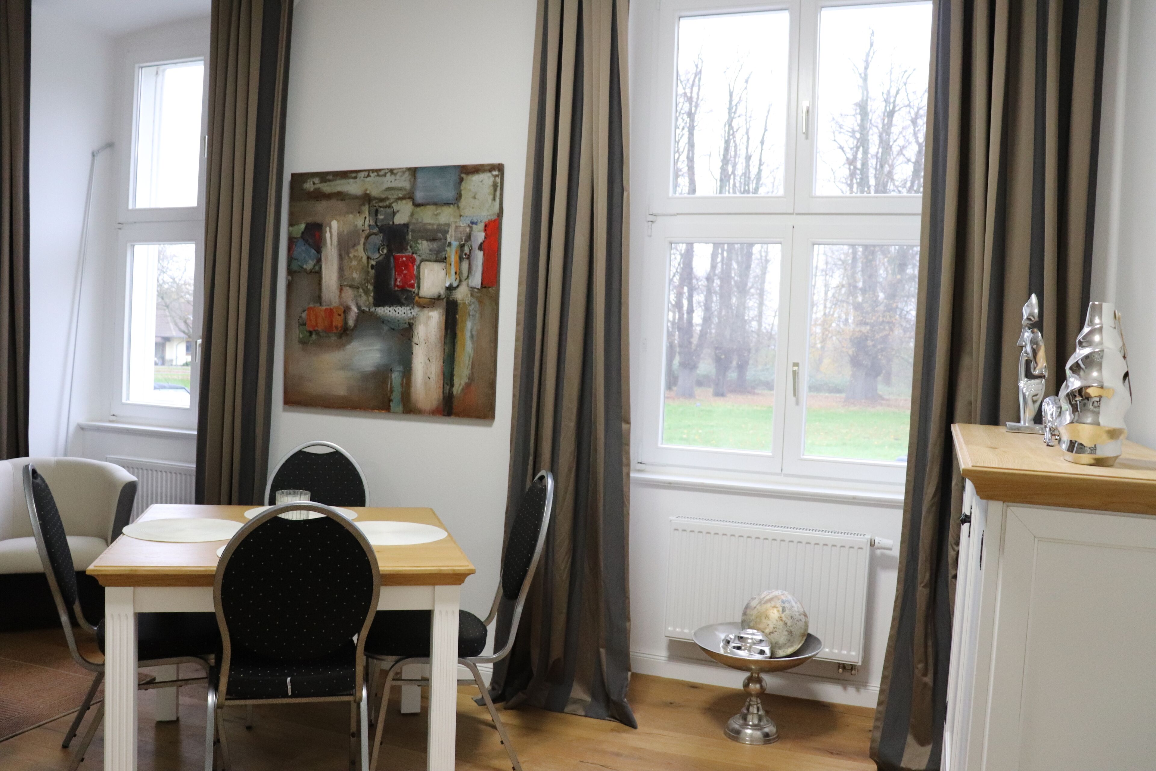 A dining area with a table, chairs, a painting on the wall, and a window with curtains.