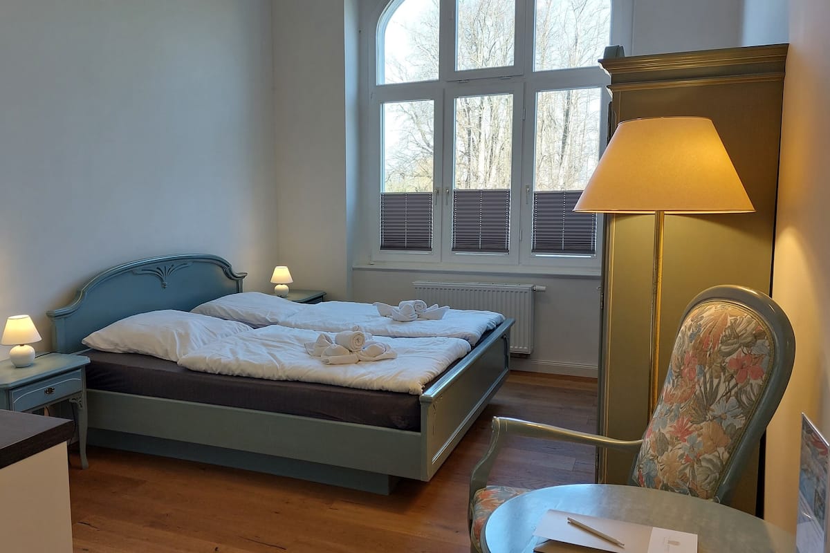 A bedroom with a bed, bedside lamps, a chair, a table, and a window with blinds.