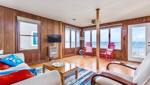 Amplia sala de estar con paredes de madera, ventanas grandes con vistas al océano, un televisor y un comedor con sillas rojas.