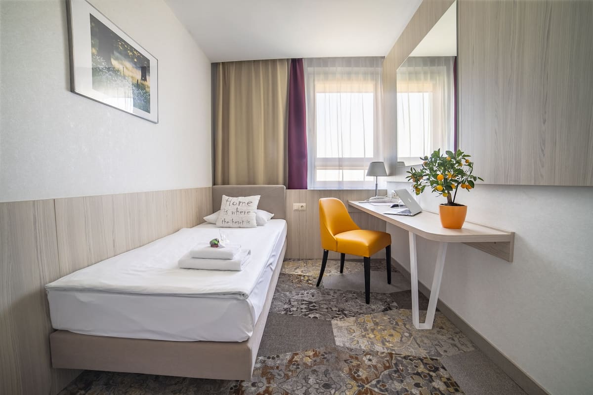 Habitación de hotel con cama, escritorio, silla, planta en maceta y ventana con cortinas.