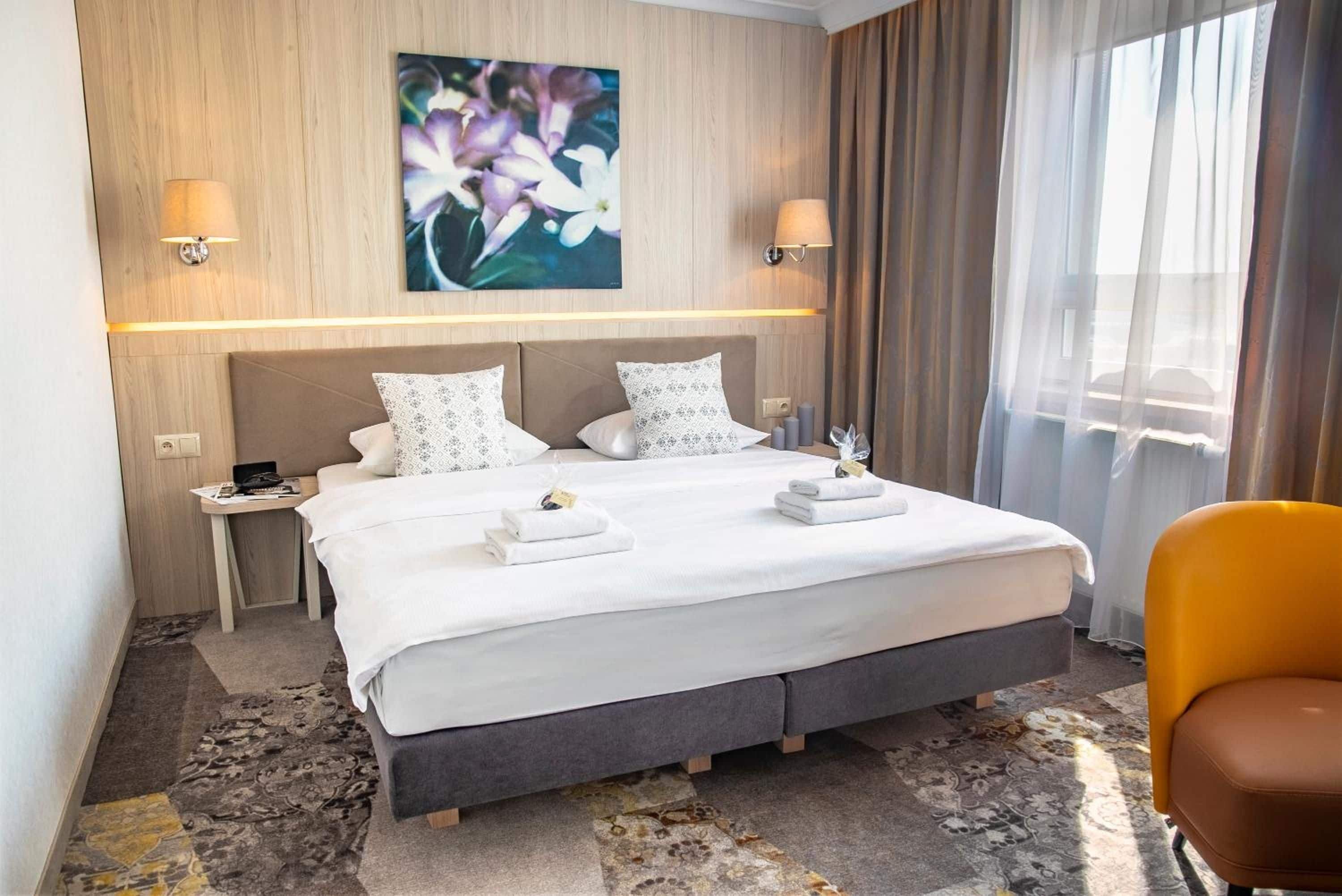 Habitación de hotel con una cama grande, dos lámparas de noche, una mesita, una silla y un ventanal con cortinas.