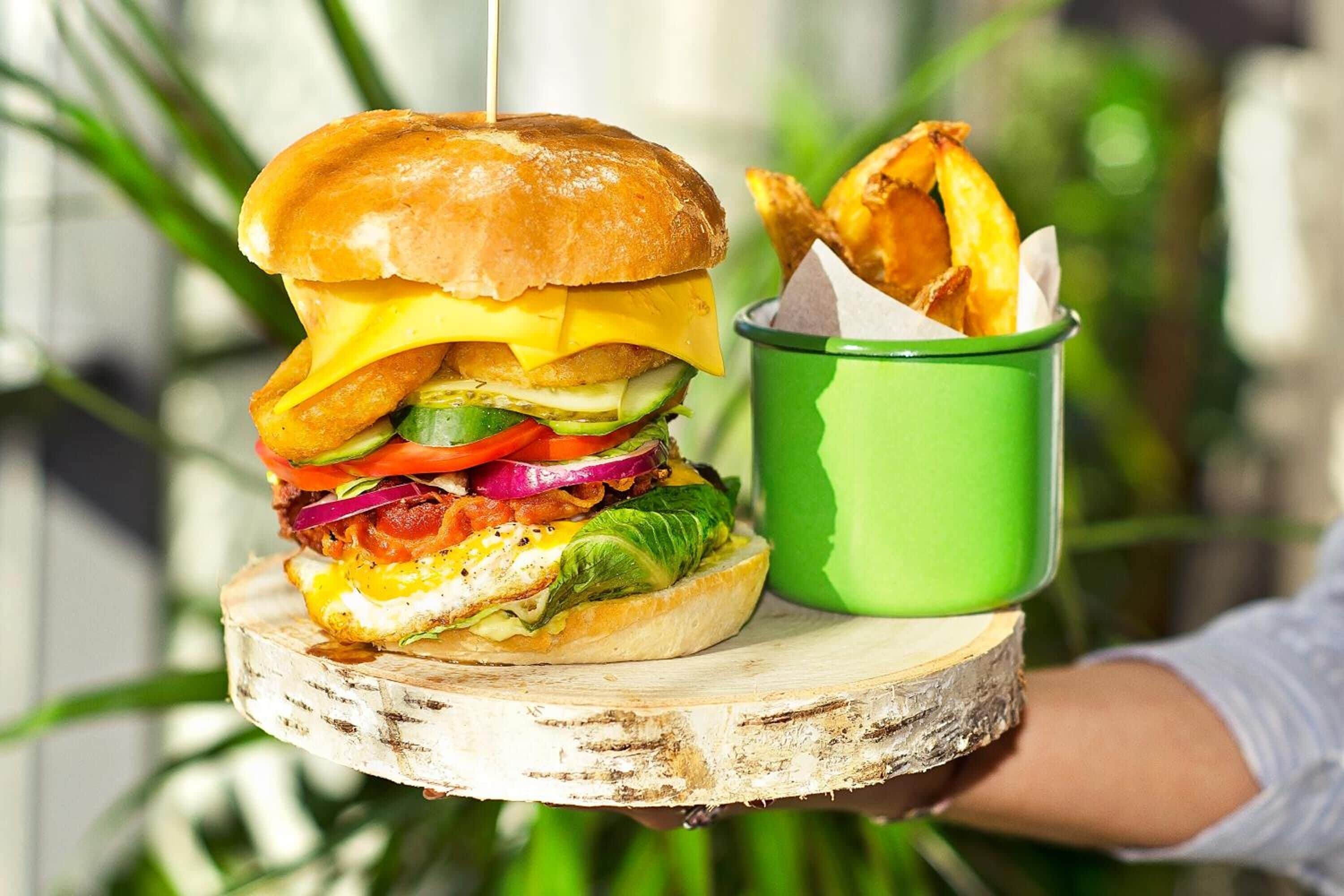 A burger with various toppings and a side of fries in a green cup, served on a wooden board.
