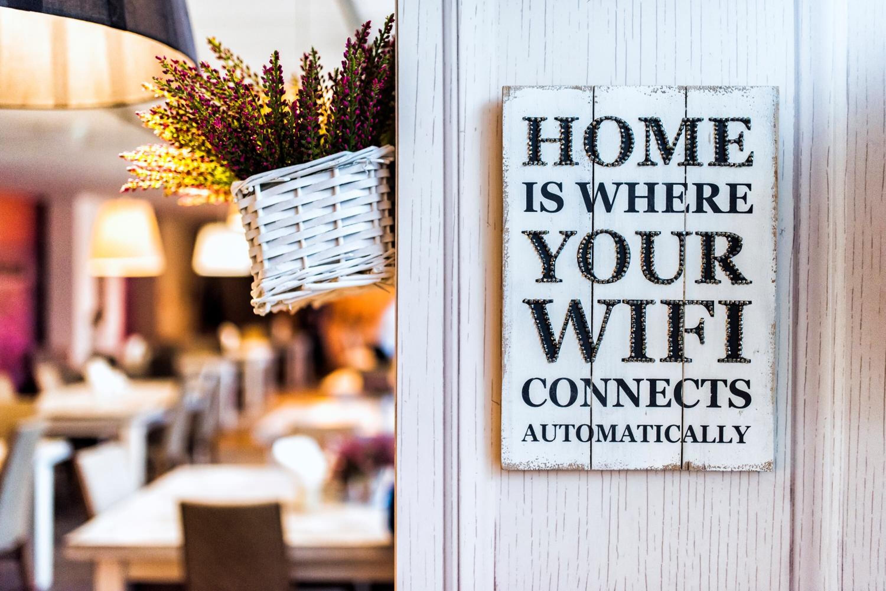 A sign that reads "HOME IS WHERE YOUR WIFI CONNECTS AUTOMATICALLY" on a white wooden wall.