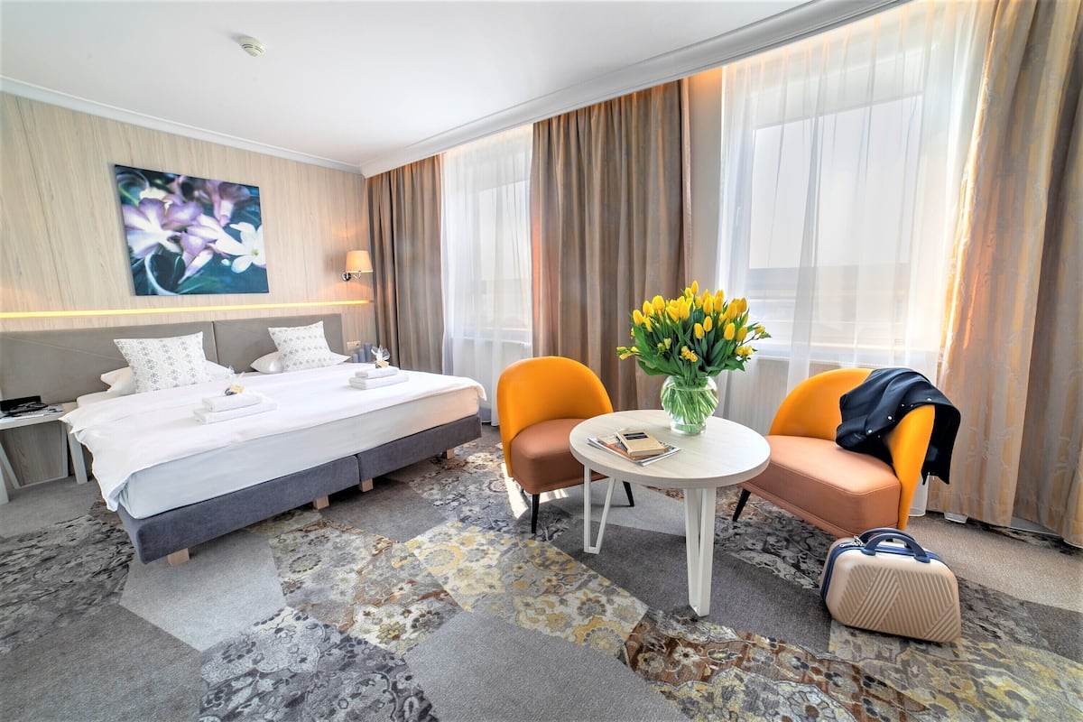 Una habitación de hotel con una cama, dos sillas, una mesita con un florero y una maleta.