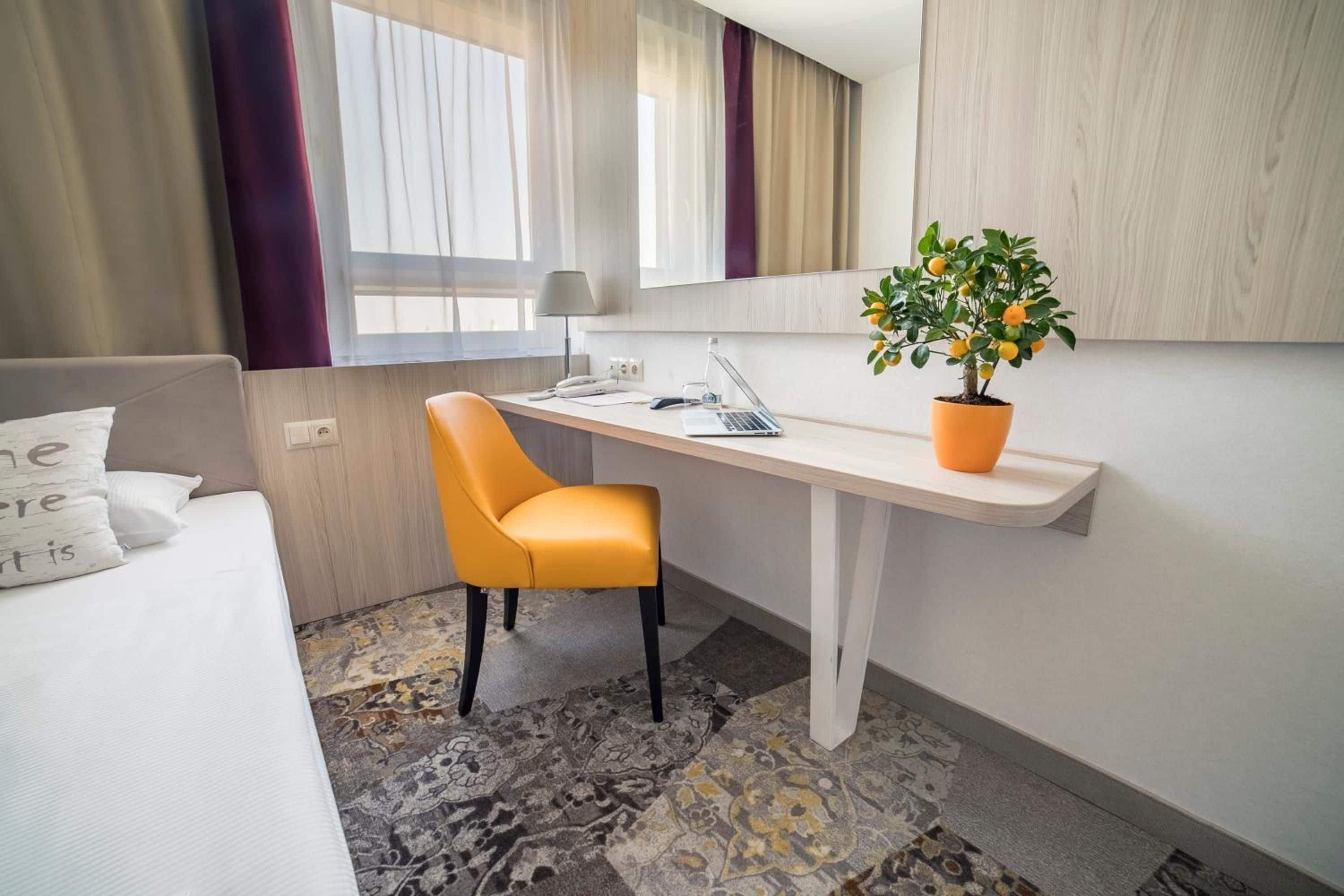 A hotel room with a bed, a desk with a laptop, a chair, a potted plant, and a window with curtains.