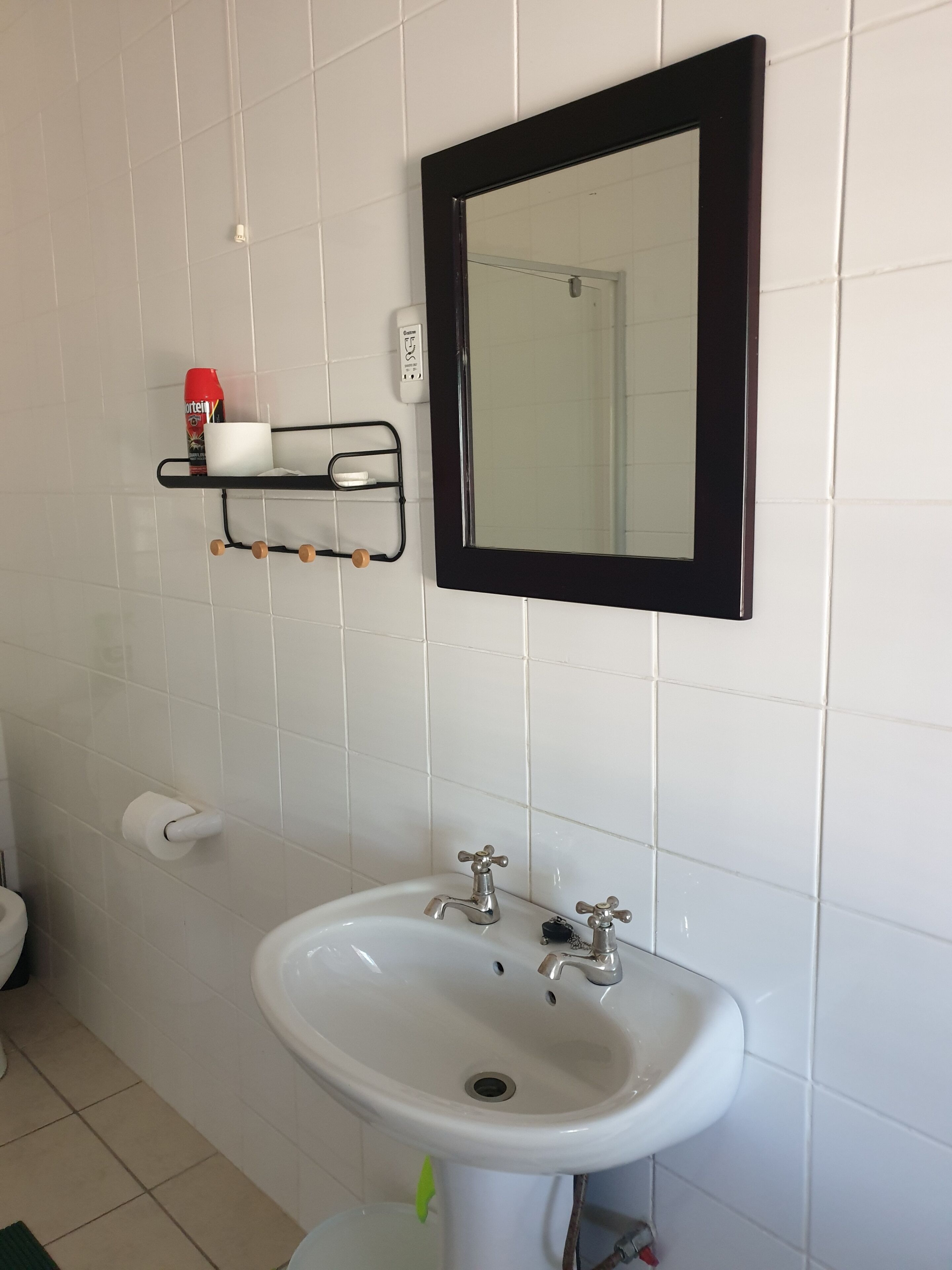 A bathroom with a white sink, two faucets, a mirror with a black frame, a toilet, and a shelf with a red can and a white cup.