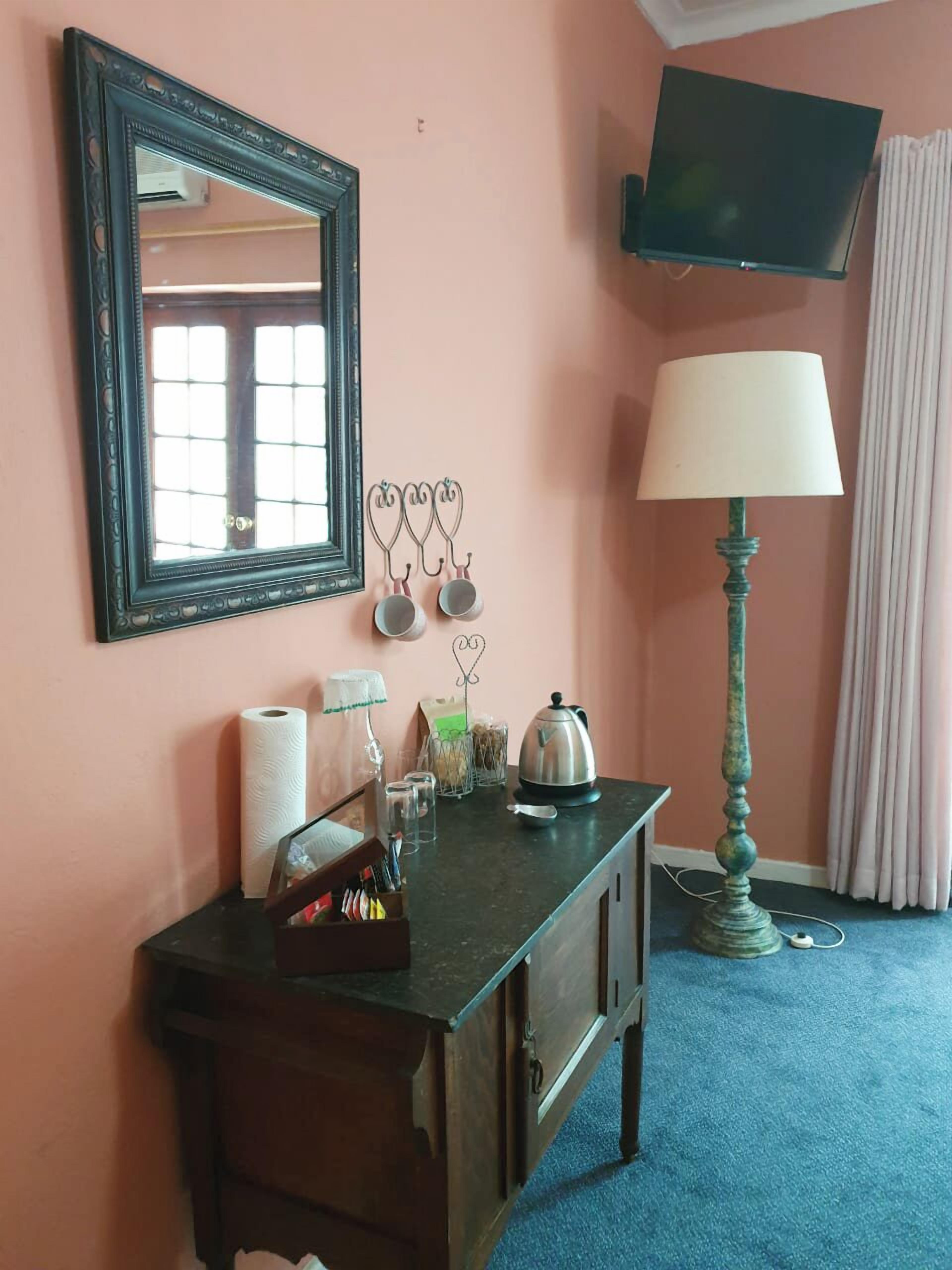 A hotel room with a wooden desk, a mirror, a TV mounted on the wall, a lamp, and a window with curtains.