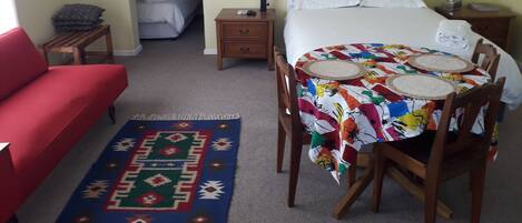 A hotel room with a bed, a red sofa, a dining table with chairs, a small bedside table, and a nightstand.