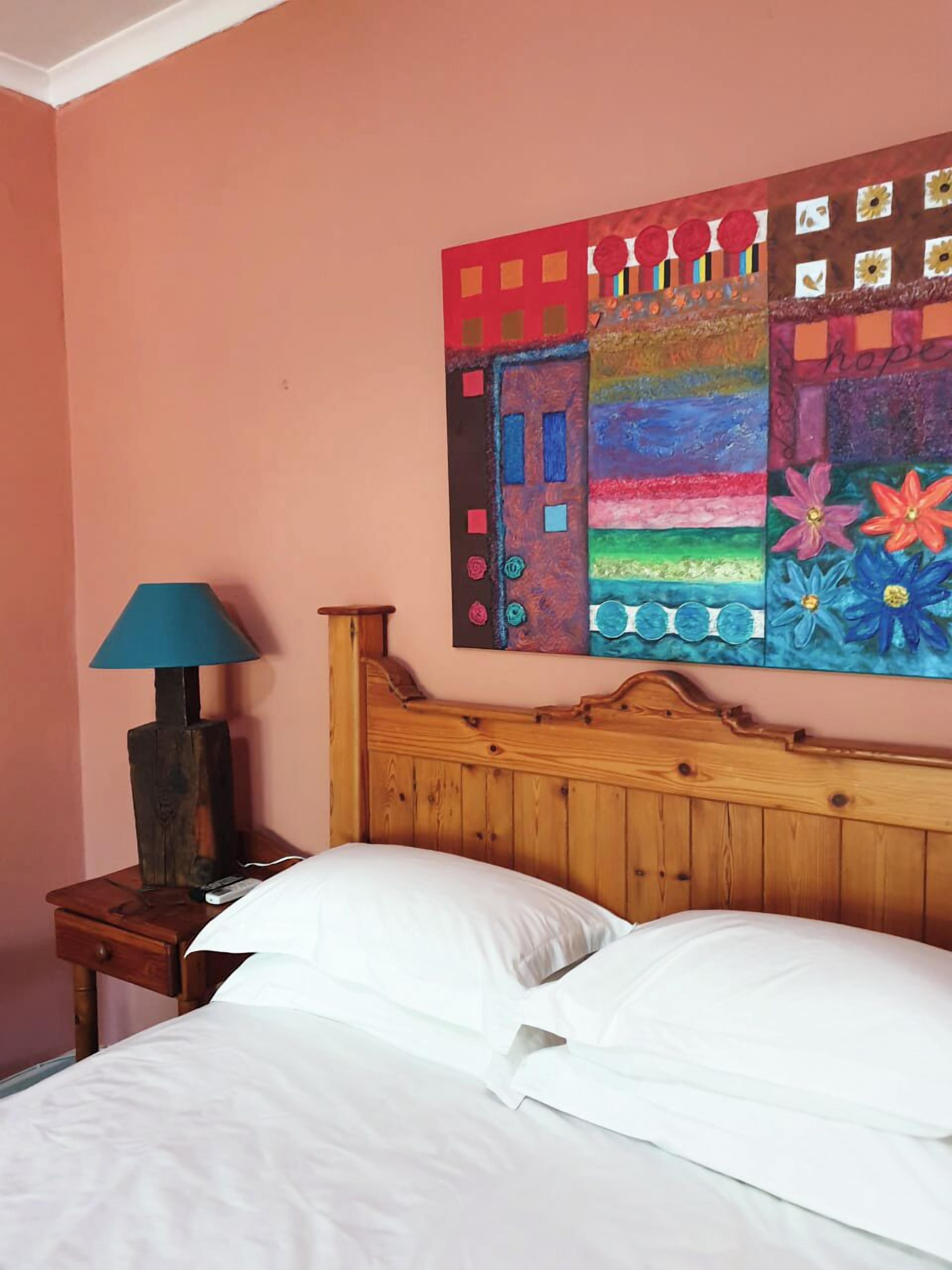 A bedroom with a wooden headboard, a bed with white linens, a nightstand with a lamp, and a colorful abstract painting on the wall.