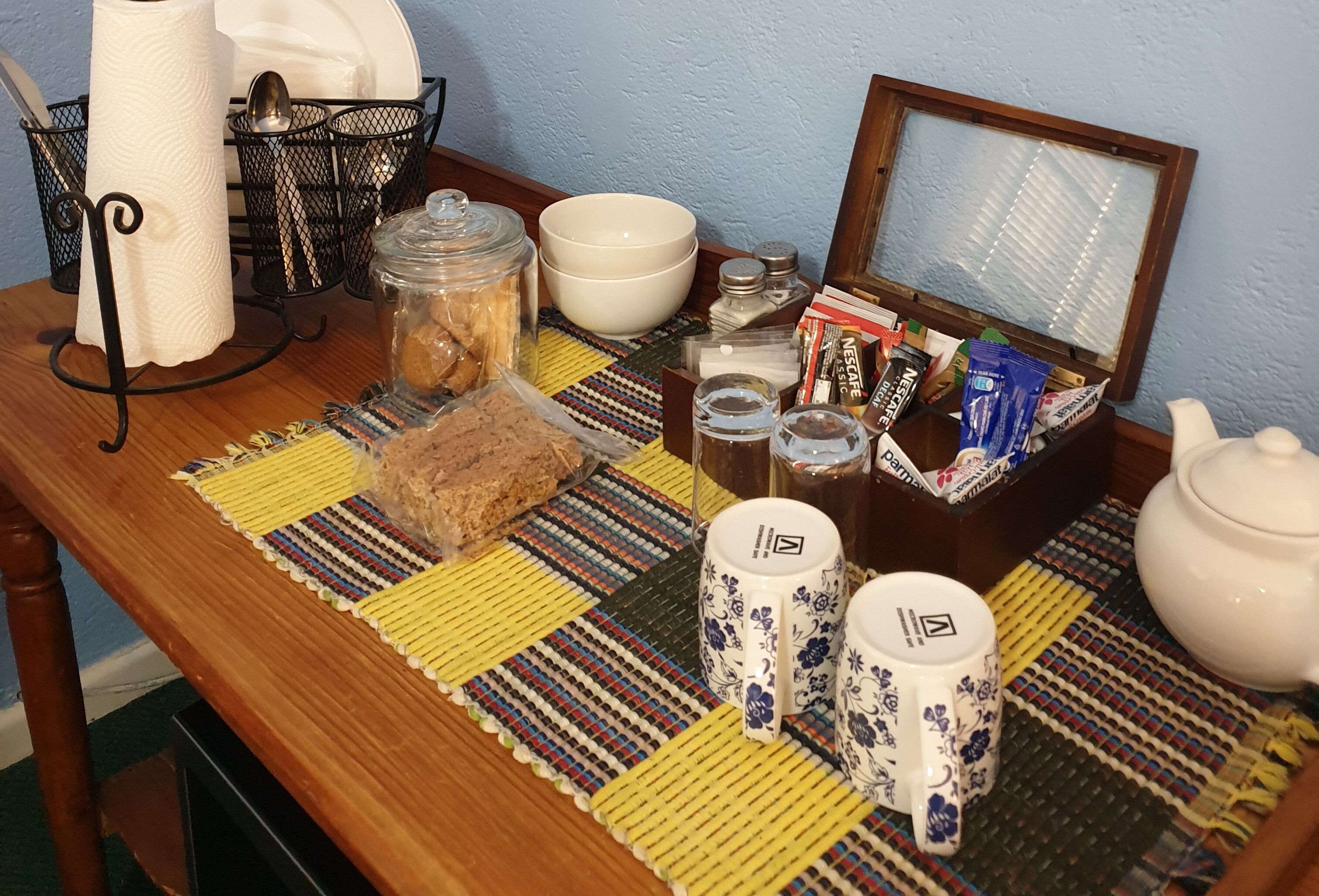A wooden table with a colorful woven placemat, a glass jar, a loaf of bread, a white bowl, a white teapot, two white cups with floral designs, a metal basket with utensils, and a framed picture.