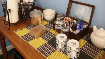 A wooden table with a colorful woven placemat, a glass jar, a loaf of bread, a white bowl, a white teapot, two white cups with floral designs, a metal basket with utensils, and a framed picture.