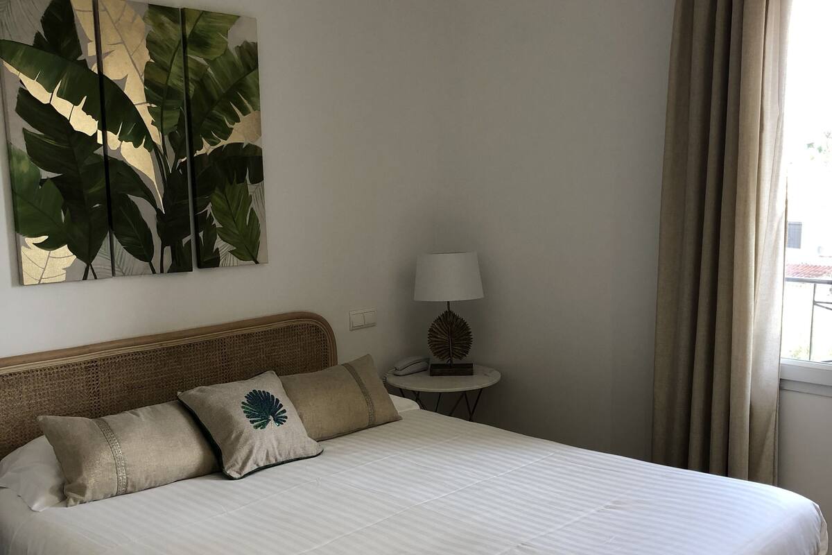 A neatly made bed with a white comforter and two pillows, a wall-mounted plant, a bedside lamp, and a window with curtains.