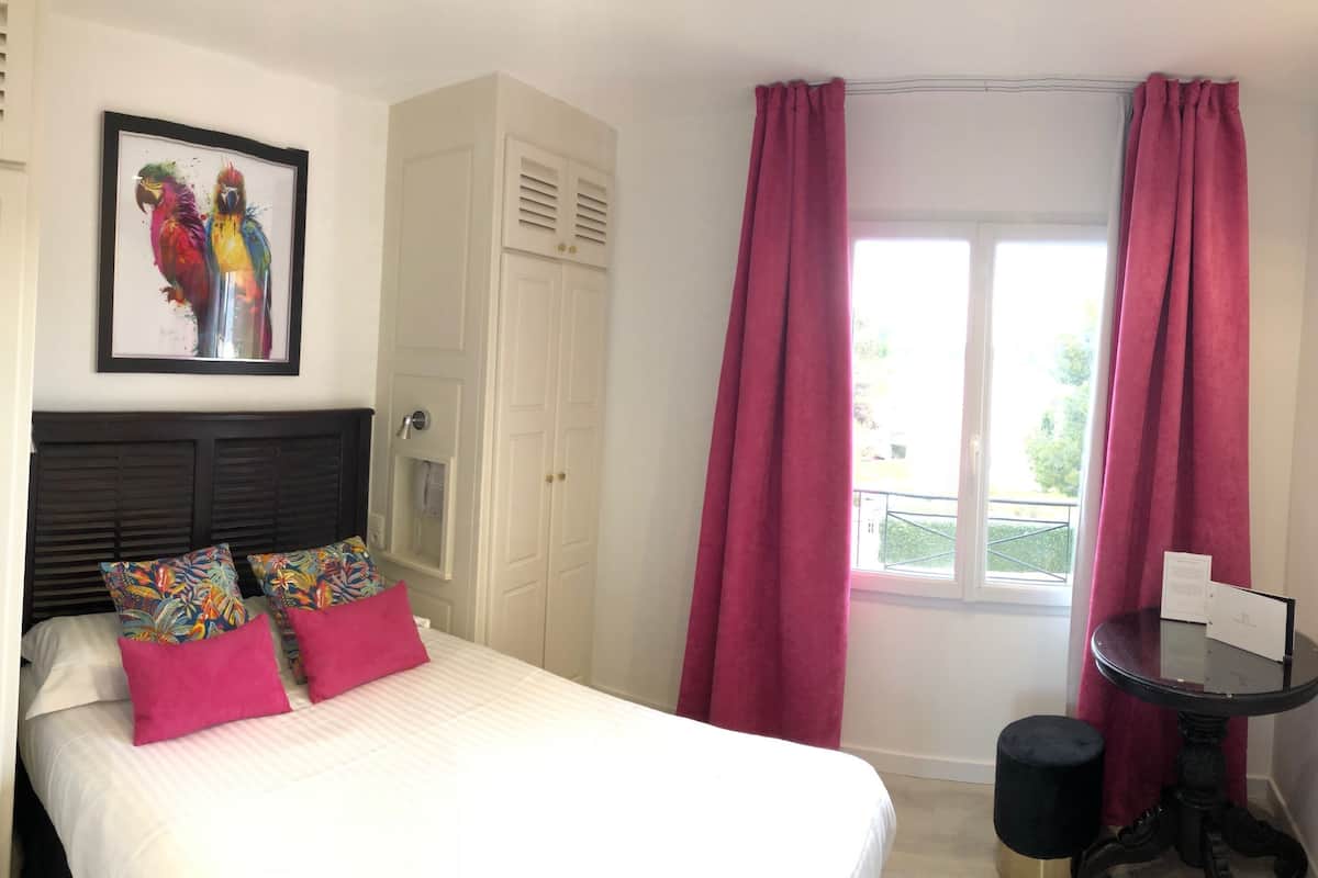 A bedroom with a bed, a framed artwork above the bed, a window with curtains, and a small table with a chair.
