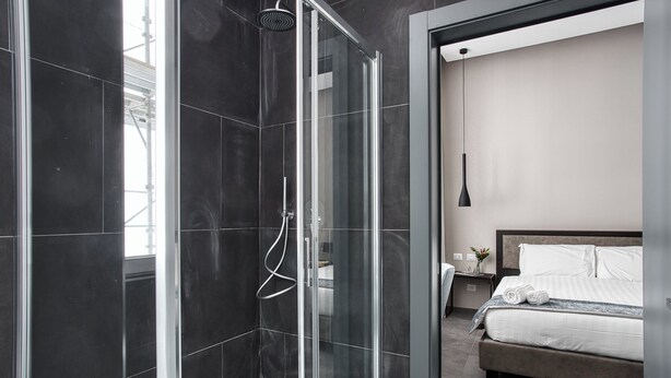A modern bathroom with a glass-enclosed shower, a large bed with white linens, and a minimalist design. - JOIVY Spadari (Milan)