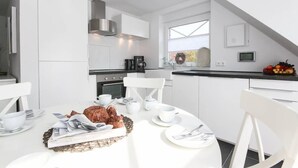 A modern kitchen with white cabinetry, a dining table set for breakfast, and a view of the outdoors through a large window. - Adams Kajüte (Krummhoern)