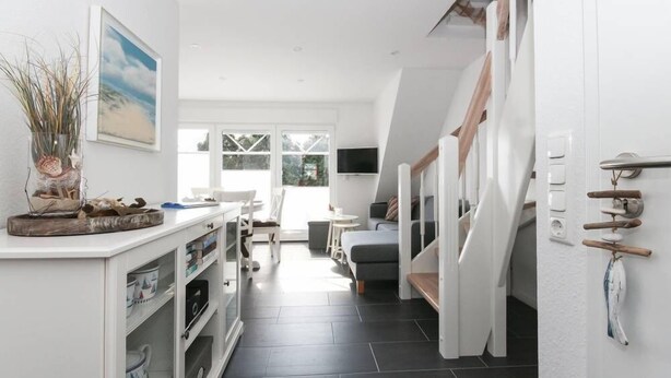 A modern, well-lit interior with a kitchen area, dining space, and a staircase leading to an upper level. - Adams Kajüte (Krummhoern)