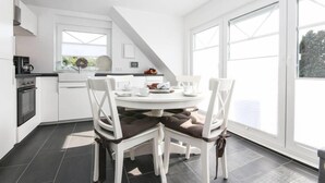 A modern kitchen with white cabinetry, a round dining table with four chairs, and large windows providing natural light. - Adams Kajüte (Krummhoern)
