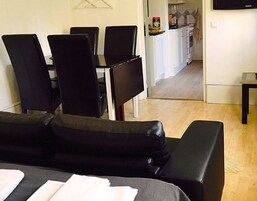 Sala de estar com sofá preto, mesa de jantar com cadeiras e área de cozinha ao fundo.