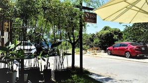 Vue de l’hôtel Oasis, pris depuis le bord de la route, avec un panneau indicateur, des plantes en pot et une voiture rouge garée à proximité.