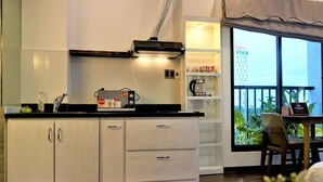 Une cuisine compacte avec des meubles blancs, un plan de travail noir et une machine à café. Une fenêtre offre une vue sur un immeuble et des plantes à l’extérieur.