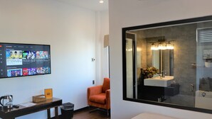 Ein modernes Hotelzimmer mit einem großen Fernseher, der einen Streaming-Dienst anzeigt, einem bequemen orangefarbenen Sessel, einem Holztisch mit einer Kaffeemaschine und einem großen Fenster mit Blick ins Bad.